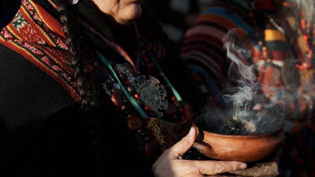 Mulher indígena em trajes tradicionais segurando uma cumbuca de onde saem fumos de incenso