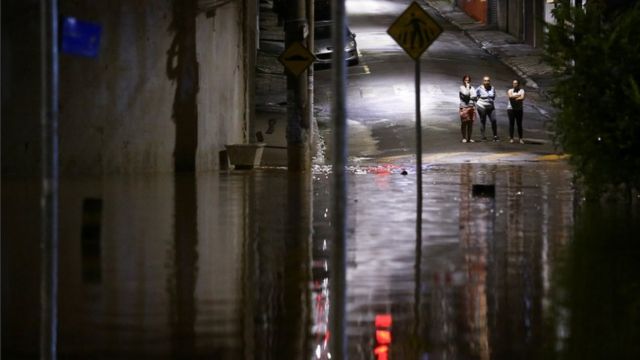 Foto noturna mostra pessoas na rua observando enchente à frente delas