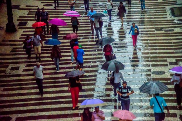 Pessoas caminhando em calçada em dia chuvoso; algumas carregam guarda-chuva