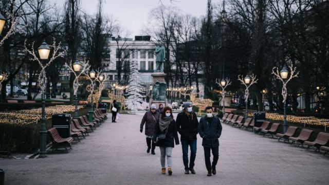 Fotografia colorida mostra jovens de máscara caminhando em uma boulevard coberta por luzinhas de Natal