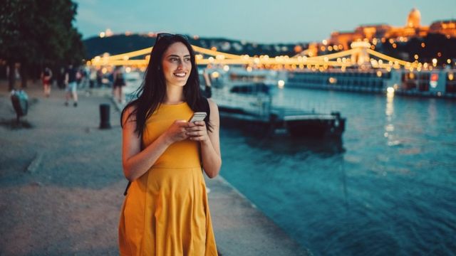 Mujer joven con un celular en sus manos. Sonríe y lleva un vestido amarillo junto al río en Budapest al atardecer