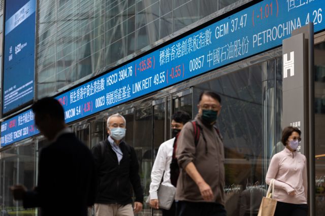 Pedestres com máscara passando em frente a painel com preços em bolsa de valores chinesa