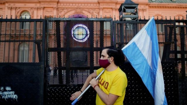 Homem de máscara segura bandeira da argentina apoiada no ombro em frente a portão da Casa Rosada