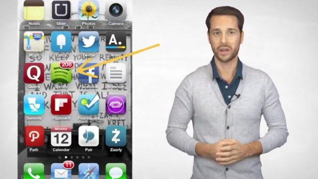 Un homme en cardigan s'adresse à la caméra - il est représenté à côté d'un téléphone portable avec des applications affichées.