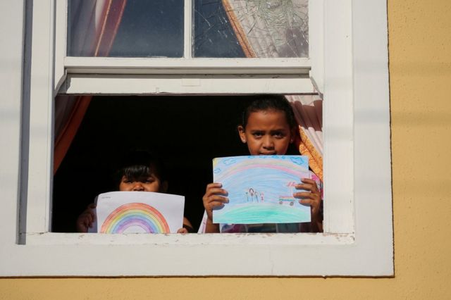 Two children stand at the window holding their drawing of what they miss most - their illustrations show rainbows and ice-cream vans.