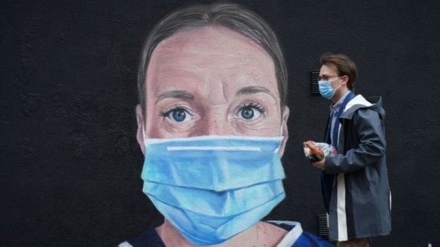 Homem de máscara caminha em frente a grafite de mulher com máscara em um muro