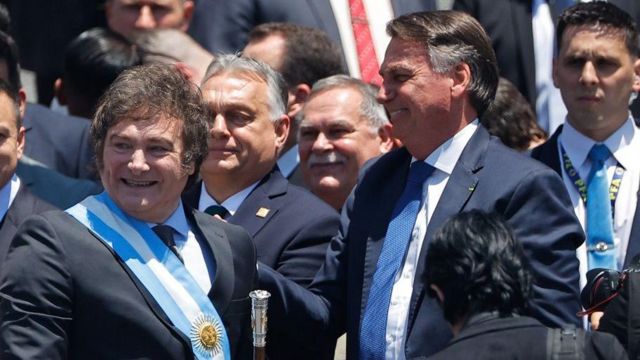 Milei e Bolsonaro sorrindo durante evento; eles estão em área externa e rodeados de outras pessoas