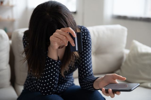 Una mujer sostiene su celular en una mano y en la otra su tarjeta de crédito.