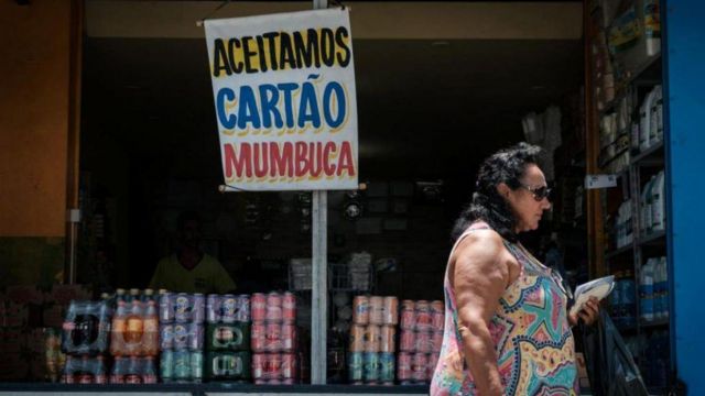 Mulher passa em frente a comércio com placa dizendo "Aceitamos cartão Mumbuca", moeda social do programa de renda básica do município de Maricá (RJ)