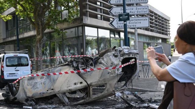 Mulher fotografa com o celular um carro incendiado e capotado