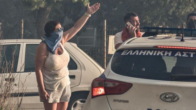  Uma mulher cobre o rosto para se proteger da fumaça, perto de carro e de outro homem que observa a situação 