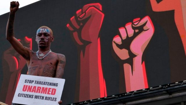 Homem com cartaz pedindo para que parem de tratar cidadãos desarmados com fuzis