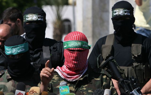 Abu Ubaida em pé diante de microfones com dois homens armados mascarados atrás dele