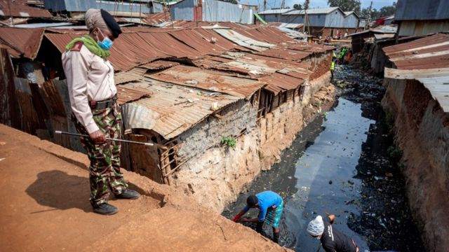 Someone looking at people cleaning an open sewer between corrugated iron houses in Kibera, Nairobi, Kenya