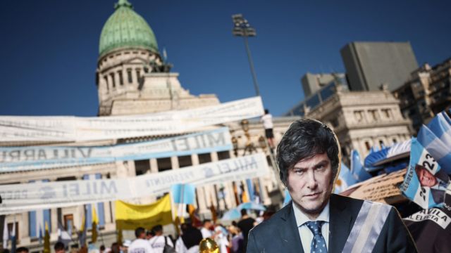 Foto colorida mostra figura de papelão com foto de Milei em frente à Casa Rosada durante posse presidencial