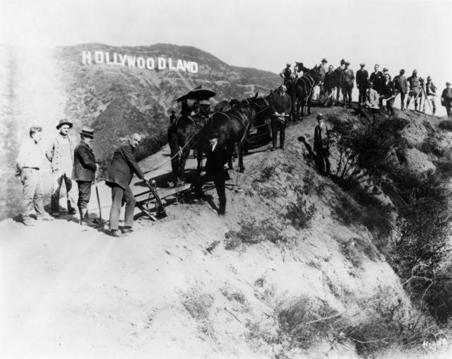 Um grupo de homens, provavelmente topógrafos e construtores que trabalham no novo conjunto habitacional conhecido como Hollywoodland, posam para um retrato abaixo da placa erguida para anunciar o local, por volta de 1925, em Los Angeles, Califórnia. (Foto de Arquivos Michael Ochs/Getty Images)