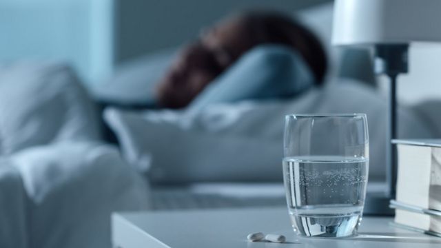 Pessoa dormindo; em primeiro plano, um copo com água e um comprimido
