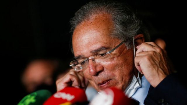 Paulo Guedes olha para baixo, diante de vários microfones de veículos da imprensa