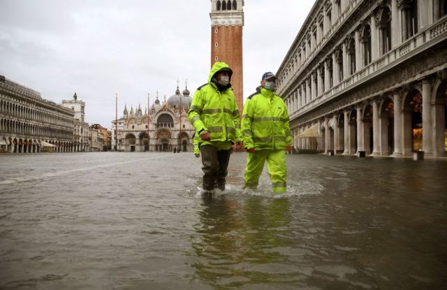 رجال يرتدون ملابس مقاومة للماء يمشون في مياه الفيضانات في ساحة سان ماركو في البندقية، إيطاليا
