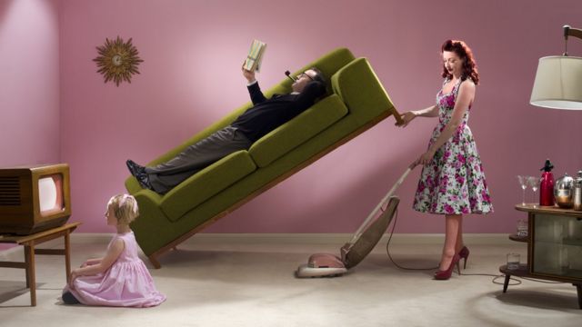 Una mujer limpiando mientras su marido está recostado en el sofá
