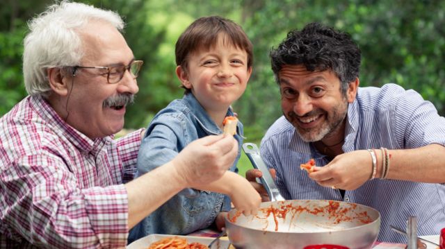 um menino e dois adultos comendo macarronada