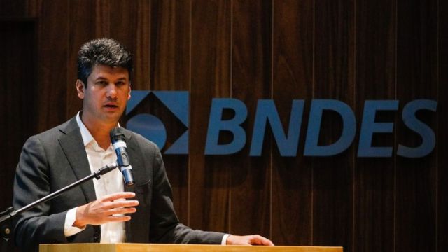Gustavo Montezano fala ao microfone em frente ao logotipo do BNDES