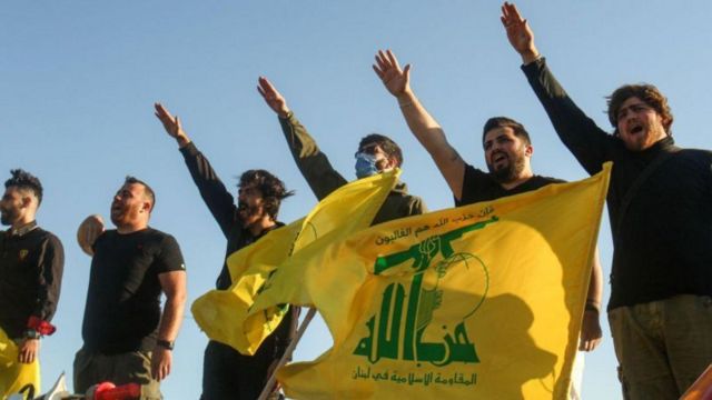 Apoiadores do Hezbollah fazendo saudação enquanto seguram bandeira do grupo