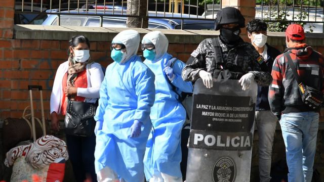Un agente espera junto a varias personas con mascarillas y material protector en Cochabamba, Bolivia