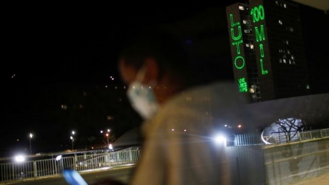 Foto à noite, mostra mulher parada em ponto de ônibus com máscara e, ao fundo, uma projeção de luz no prédio do Congresso dizendo: Luto 100 mil