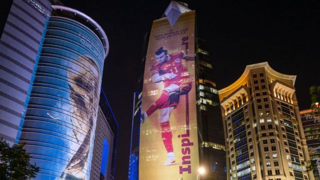 Fotografia colorida mostra arranha-céus em Doha com imagens dde um jogador de futebol e da taça da copa projetadas nas fachadas