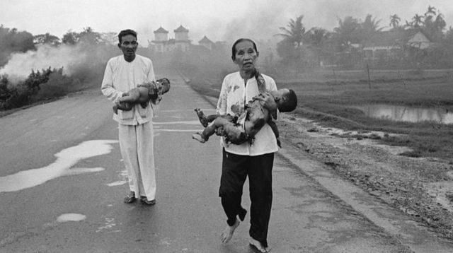 Cette photo en noir et blanc montre un homme et une femme marchant dans une rue piégée par la fumée. Chacune de ces personnes porte un bébé.