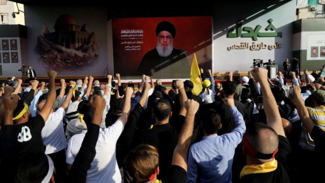 Pessoas com punhos erguidos em frente a uma imagem do líder do Hezbollah, Hassan Nasrallah