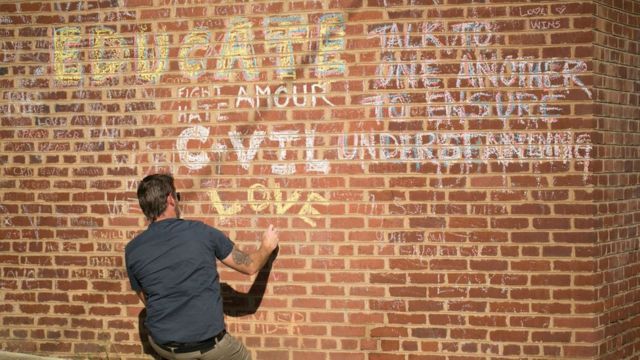 Homem agachado de costas escrevendo em parede na qual se lê mensagens que pedem compaixão 