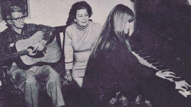 Em foto preto e branco, Rita aparece tocando piano, observada pelos pais