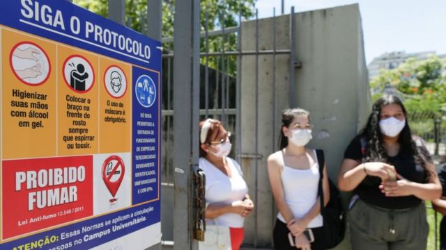 Três mulheres de máscara em dia de sol ao lado de placa com protocolo contra o coronavírus na porta de local de prova do Enem