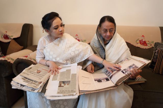Fotografia colorida mostra uma mulher jovem com roupas brancas do Bangladesh sentada no sofá ao lado de uma senhora idosa de óculos, ambos observam fotografias e recortes de jornal