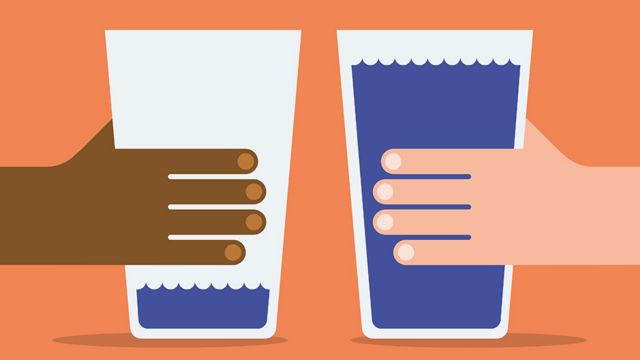 A white person's hand holds a glass full of water and a brown person's hand holds a nearly empty glass of water.