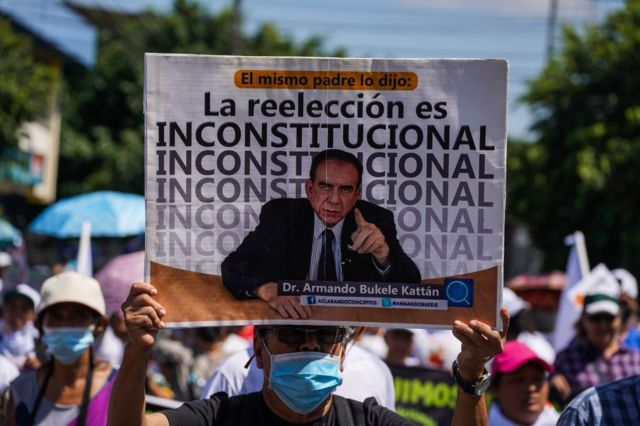Manifestante contra la candidatura presidencial de Bukele sostiene un cartel que señala que la reelección presidencial continua en El Salvador es inconstitucional.