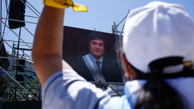 Foto colorida mostra apoiador(a) de Milei com camiseta da argentina, de costas, em frente a um telão com foto de Milei tomando posse