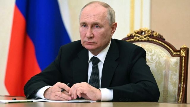 Putin sentado em frente a mesa, com olhar sério e bandeira da Rússia ao fundo