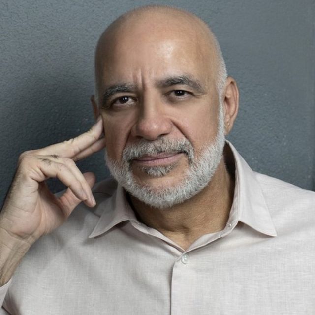 Jessé Souza, um homem negro de barca branca, de meia idade, com rosto apoiado na mão e olhar pensativo, sobre um fundo cinza