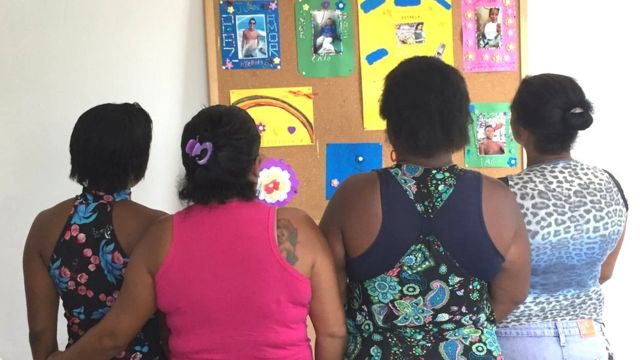 Grupo de mães olha para quadro com fotos de familiares