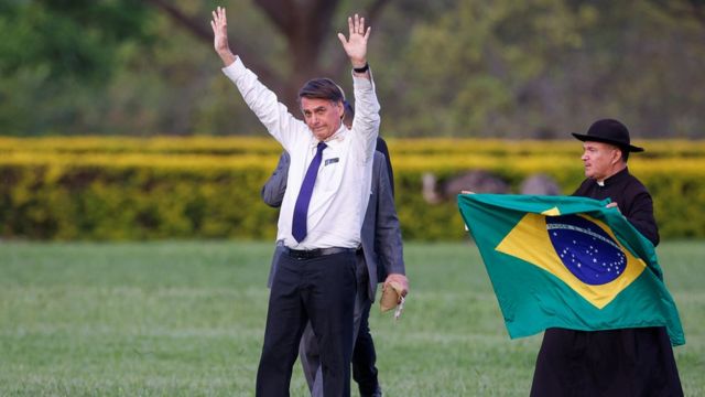 Bolsonaro em jardim, acenando em direção ao alto; ao lado dele, estão seguranças e um homem segurando bandeira do Brasil