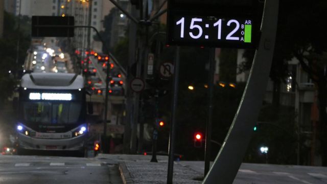 Via de São Paulo repleta de veículos, com relógio de rua marcando 15h12 e luminosidade fraca