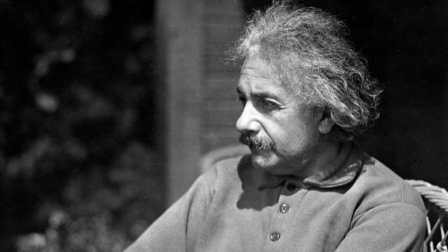 Einstein sentado vistiendo ropa informal y con la mirada perdida.