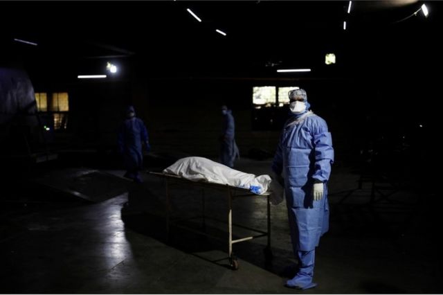 Un trabajador de la salud, vestido en traje de protección personal, espera frente al cuerpo de un hombre que murió de covid-19, antes de su cremación en Nueva Delhi, India, 21 de abril de 2021