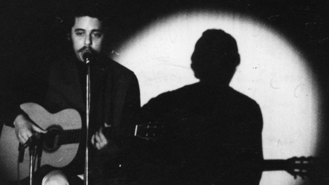 Chico Buarque no palco, sentado com violão, em foto preto e branca