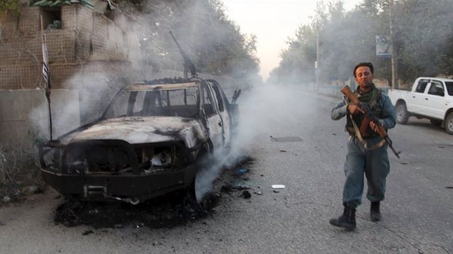 An Afghan policeman next to a burning vehicle, with a gun and the Taliban flag attached, in the city of Kunduz