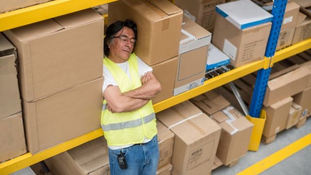 Un homme s'est endormi debout, il se repose sur une étagère pleine de cartons bruns.