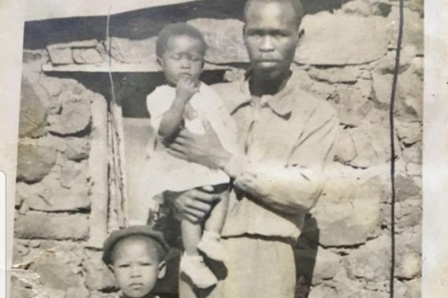 Agnes e seu pai, com sua irmã mais nova no colo, em frente à casa onde ela nasceu